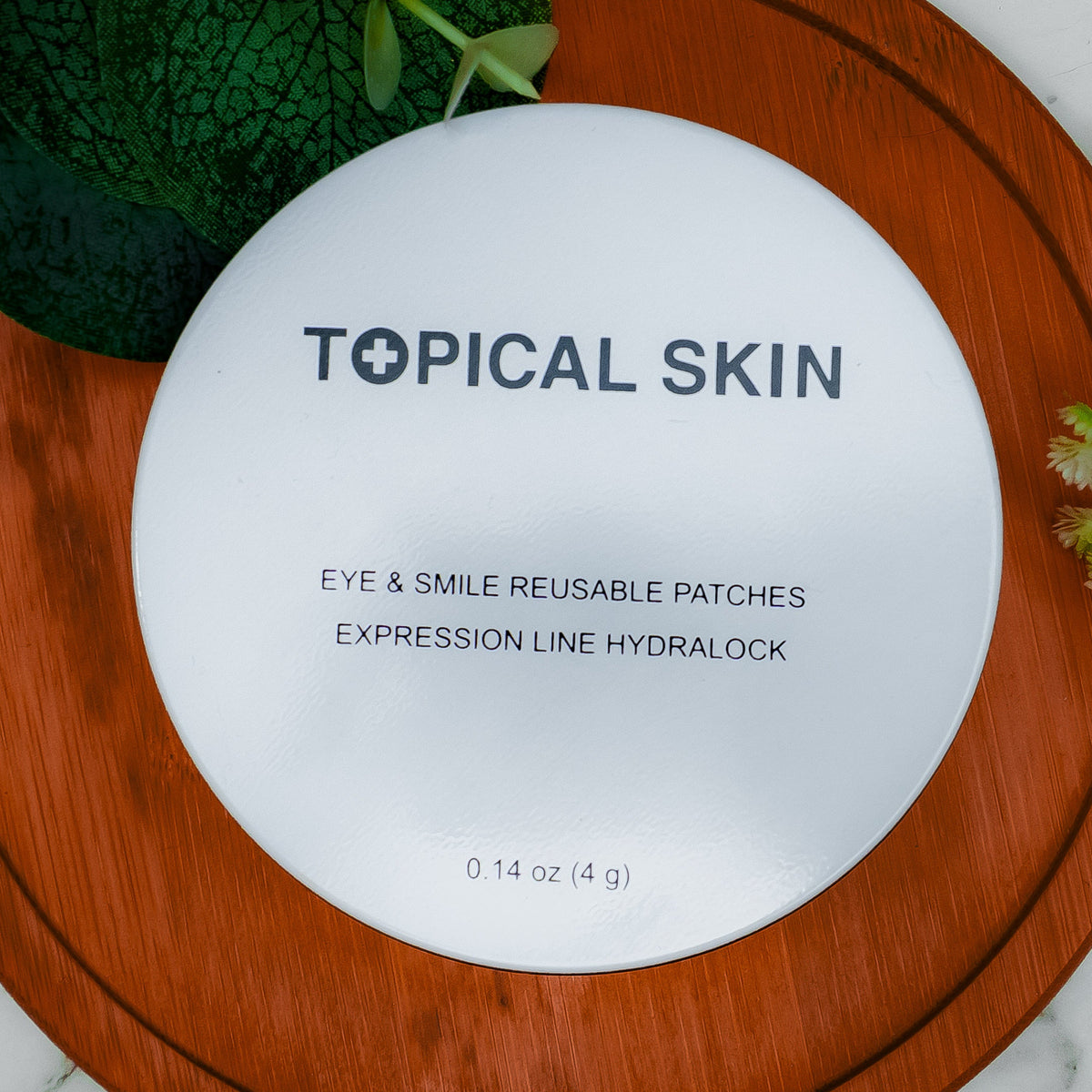 A round white cosmetic compact labeled 'TOPICAL SKIN' with text reading 'EYE & SMILE REUSABLE PATCHES' and 'EXPRESSION LINE HYDRALOCK,' placed on a round wooden board over a white marble surface with green leaves and small flowers around it.