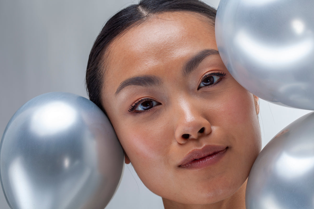 Woman’s face nestled between shiny gray and white balloons, eyes looking toward camera, soft studio lighting.