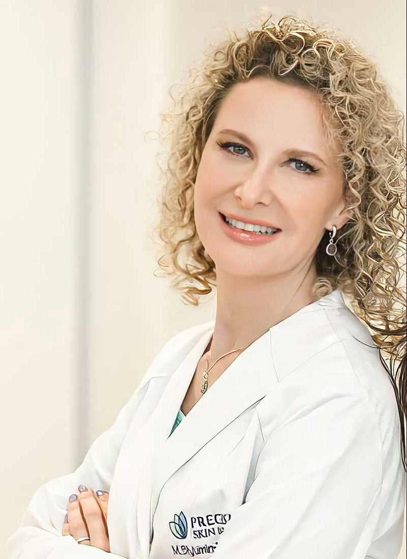 A woman with curly blonde hair in a white lab coat smiles in a softly lit studio portrait.