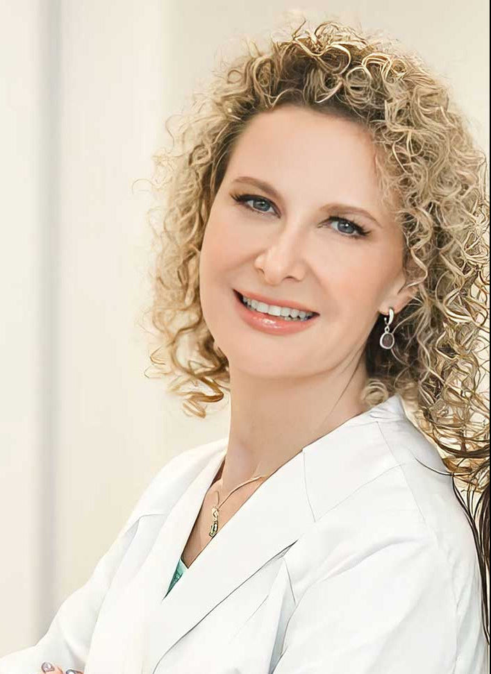 A woman with curly blonde hair in a white lab coat smiles in a softly lit studio portrait.