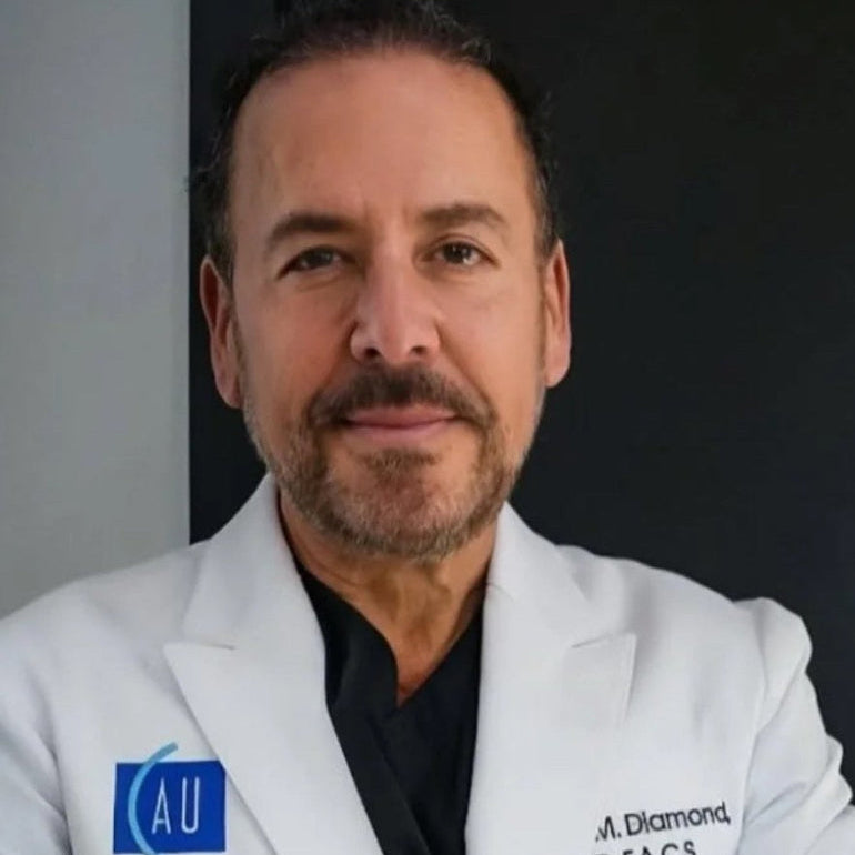 Doctor in a white lab coat with printed logos and a dark shirt, arms crossed in a studio portrait.