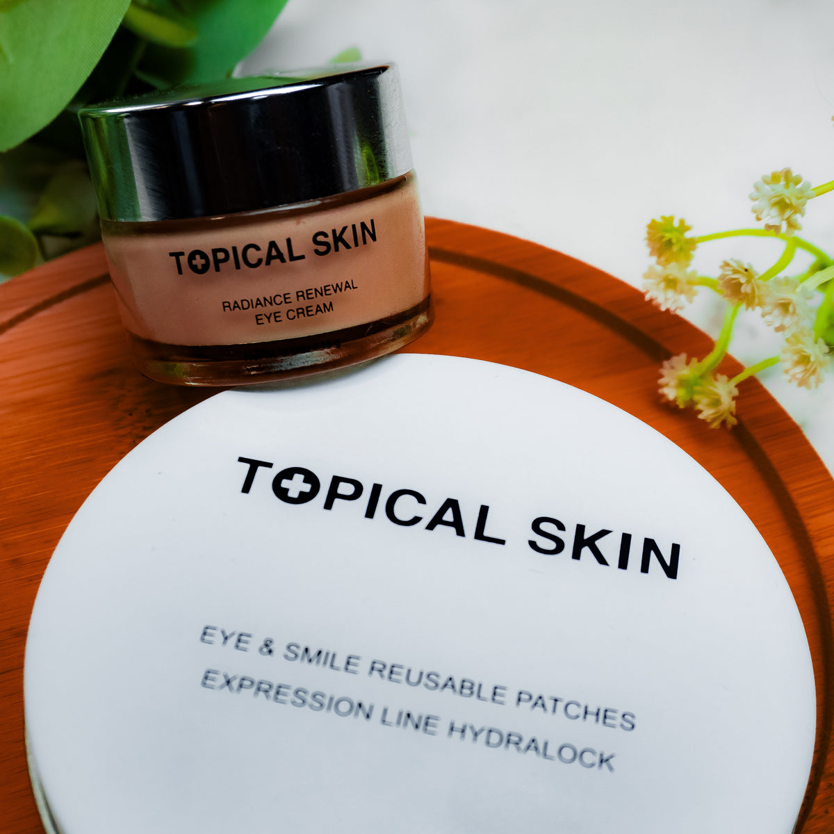 Topical Skin eye cream jar and reusable eye patches packaging arranged on a round wooden tray with green leaves and small flowers on a light marble surface.