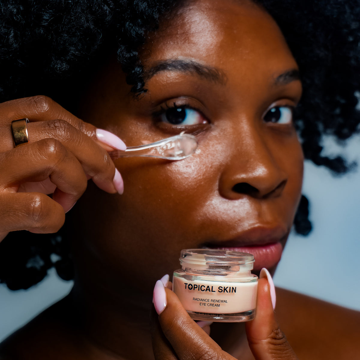 A close-up lifestyle shot of a woman applying a clear gel from a small jar labeled 'TOPICAL SKIN' to her under-eye area. She is holding the jar in one hand and using a spatula with the other, set against a solid light gray background.