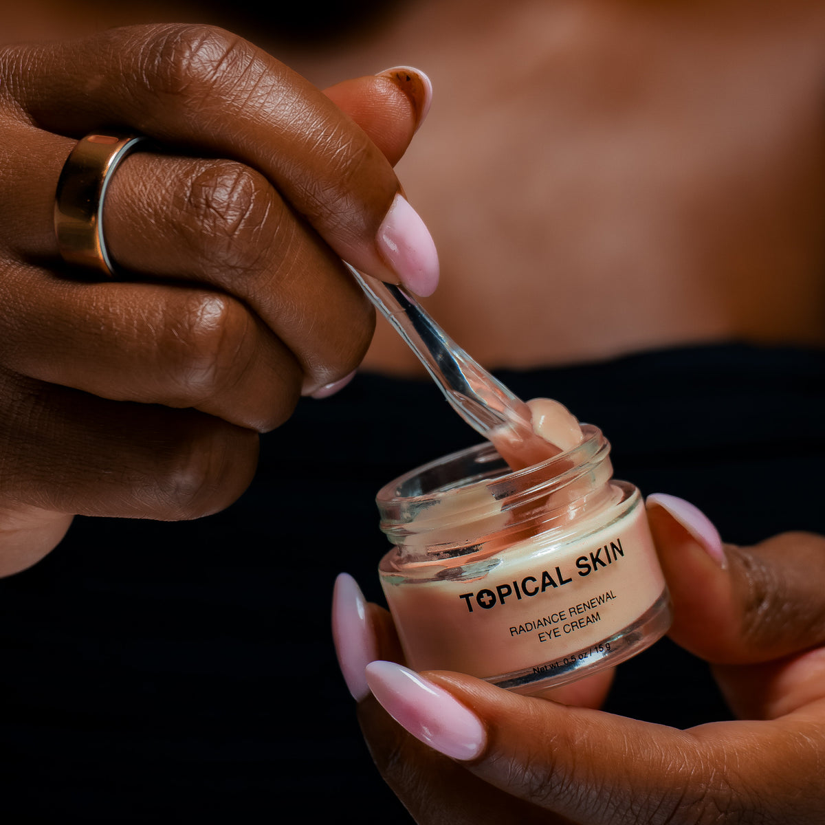 A close-up of a person holding an open jar of TOPICAL SKIN Radiant Reviver Eye Cream and scooping cream with a clear spatula. The person has dark skin, pink manicured nails, and a gold ring on one finger, wearing a black top against a dark background.