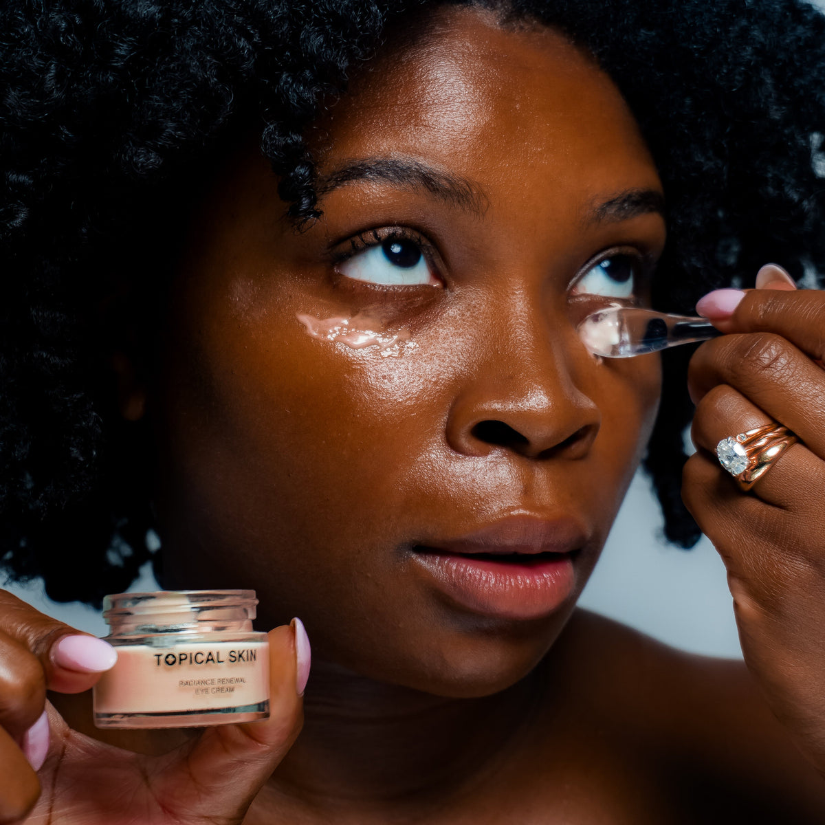 A close-up lifestyle shot of a woman applying under-eye cream from a small jar labeled TOPICAL SKIN. She is holding the open jar in one hand and using a small spatula to apply the cream to her other eye, with her other hand holding a ring nearby.