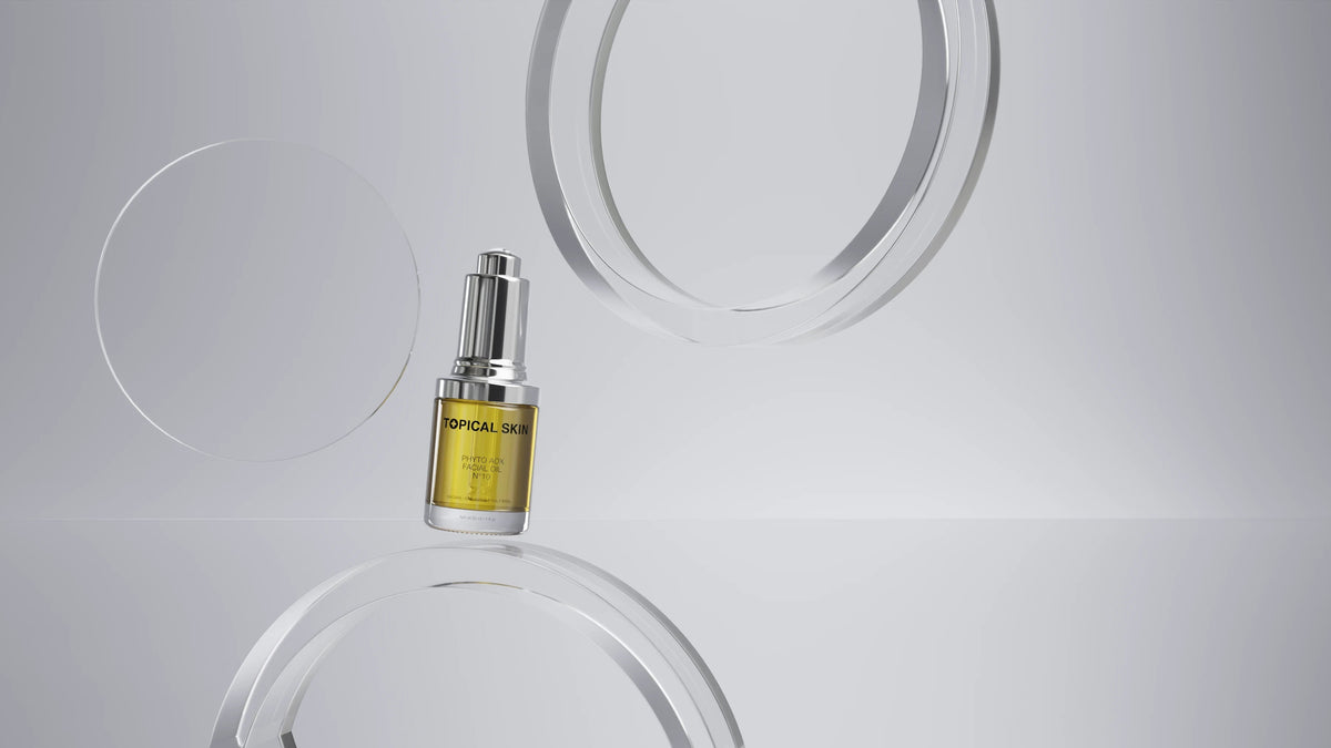 Cosmetic serum bottle with metallic cap and amber liquid, centered between smooth white circular shapes on grey.