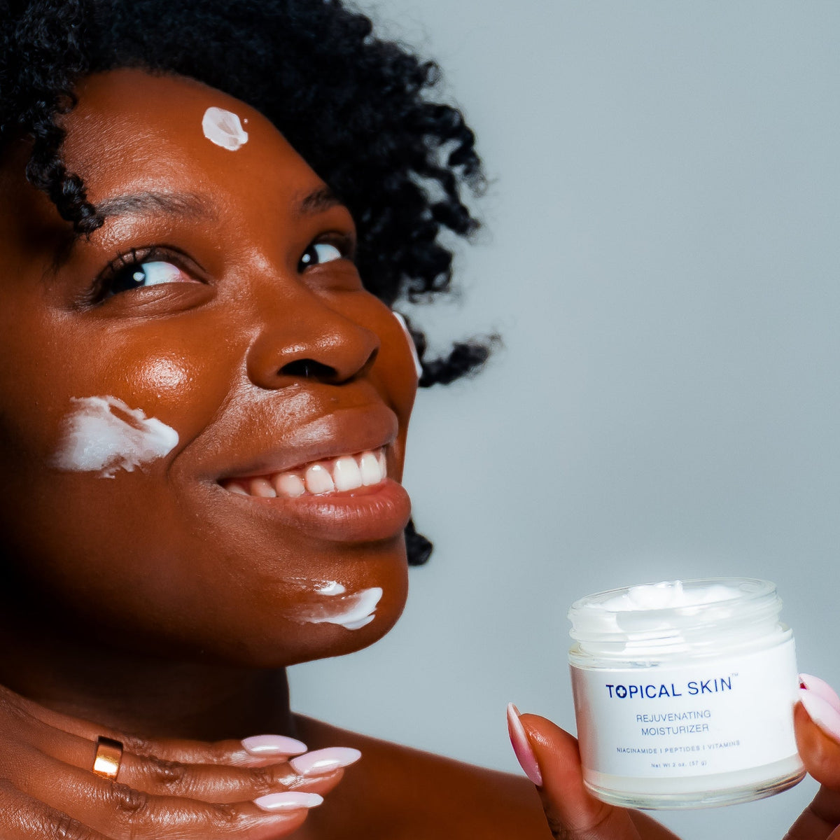 A smiling woman with dark curly hair applies white cream to her face while holding a small white jar labeled 'TOPICAL SKIN' against a plain light gray background.