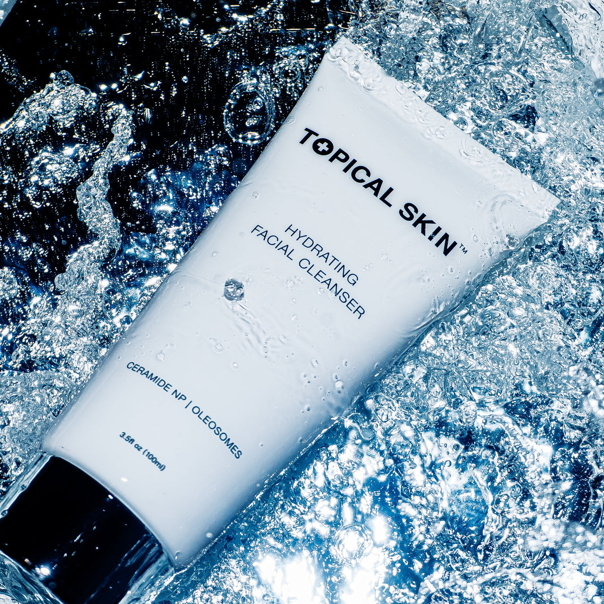 A white tube of TOPICAL SKIN Hydrating Facial Cleanser with a silver cap, surrounded by splashing water droplets and ice against a dark background.