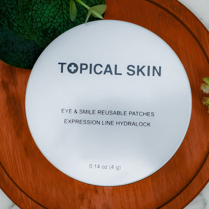 A round white cosmetic compact labeled 'TOPICAL SKIN' with text reading 'EYE & SMILE REUSABLE PATCHES' and 'EXPRESSION LINE HYDRALOCK,' placed on a round wooden board over a white marble surface with green leaves and small flowers around it.