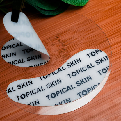 A close-up of a crescent-shaped cosmetic eye patches by TOPICAL SKIN clean dermaceuticals placed on a wooden surface, with a white backing peel partially lifted. The shield is printed with the brand name 'TOPICAL SKIN' repeated in black text.