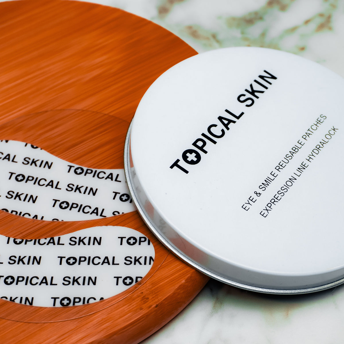 A round white metal tin of TOPICAL SKIN expression line filler sits on a white marble surface next to a wooden tray holding two white eye-shaped patch sheets with black text.