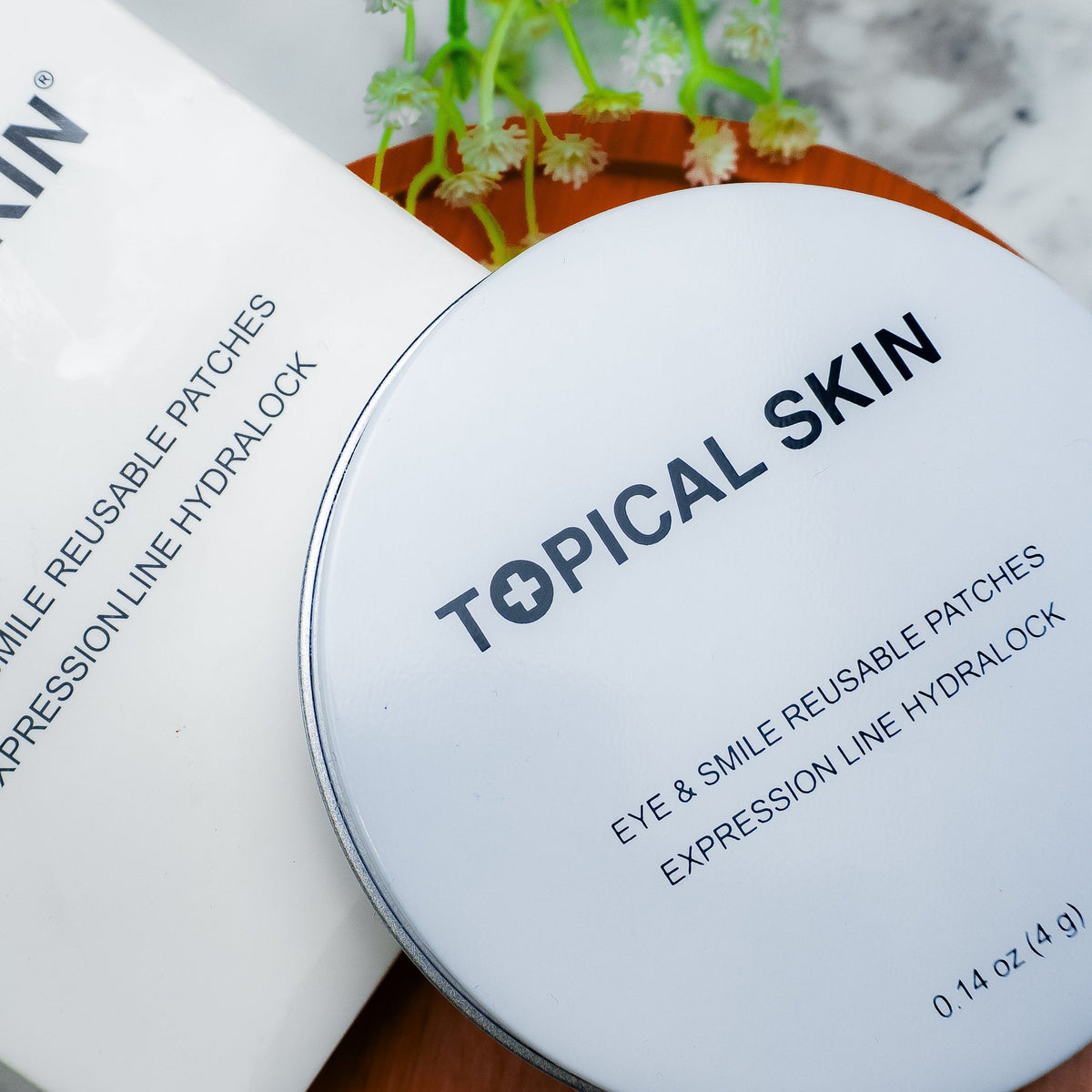 A round white lid with black text reading 'TOPICAL SKIN' and 'EYE & SMILE REUSABLE PATCHES EXPRESSION LINE HYDROLOCK' placed on top of a white product box. The scene is styled on a marble surface with a small plant in the background.